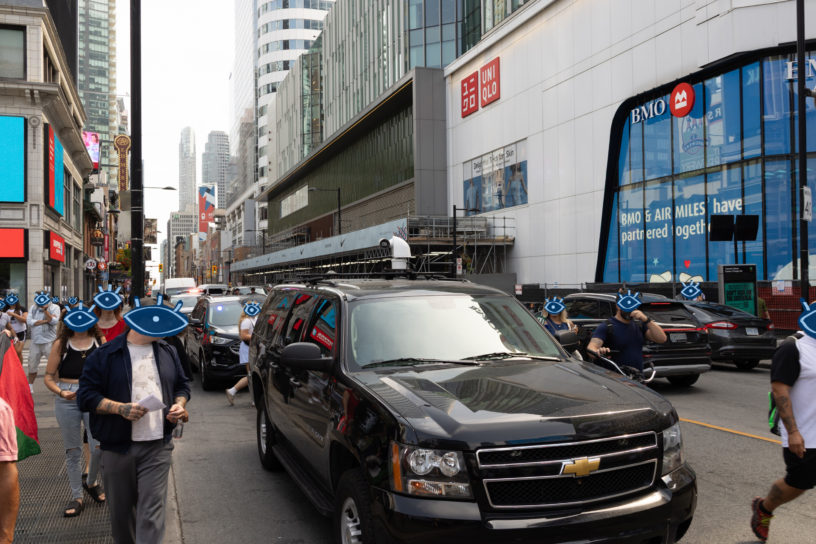 Photo of a busy street with a facial recognition camera on top of a black SUV, there are illustrations of blue eyes over every persons face.