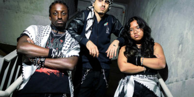 Photo of three students posing in a concrete stairwell dressed in black alternative-styled outfits. All three people are wearing black and white keffiyehs.