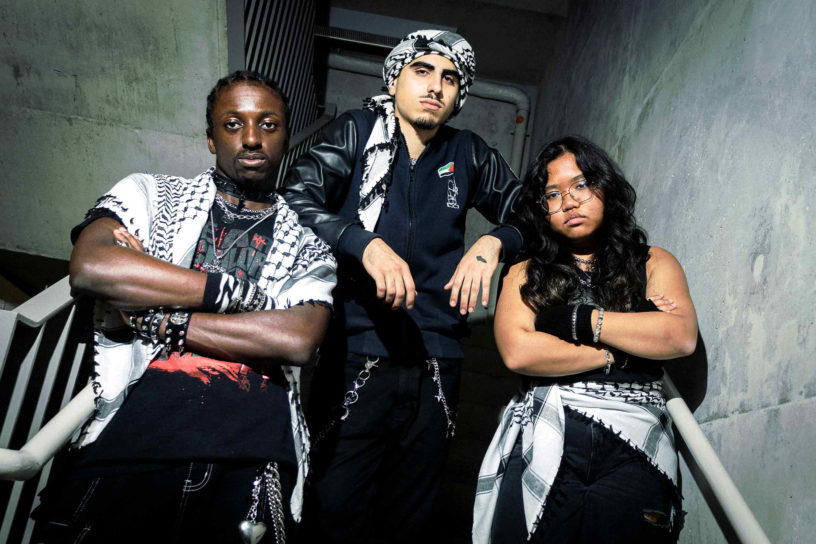Photo of three students posing in a concrete stairwell dressed in black alternative-styled outfits. All three people are wearing black and white keffiyehs.