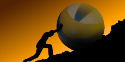A person pushing a large volleyball up a black rocky hill with an orange background.