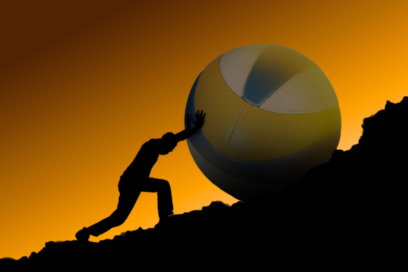 A person pushing a large volleyball up a black rocky hill with an orange background.