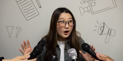 Image of a person shocked with their hands up as five microphones are pointed at their face. There are small doodles of a notepad and pencil, a video camera, a megaphone and a exclamation and question mark.