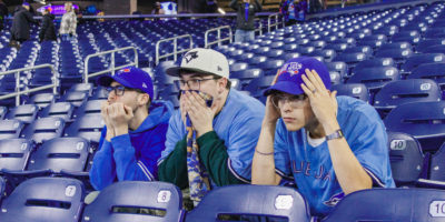Three people sitting on blue seats in Blue Jays gear looking nervously forward.