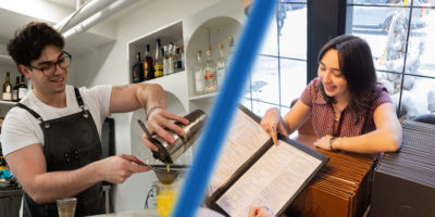 Composition of two images; on the left a bartender pours a drink and on the right a host points at the menu with guests.