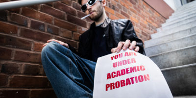 Photo of a man sitting on a staircase with a cigarette in his mouth, and holding a paper which reads "YOU ARE UNDER ACADEMIC PROBATION" in bold red text.