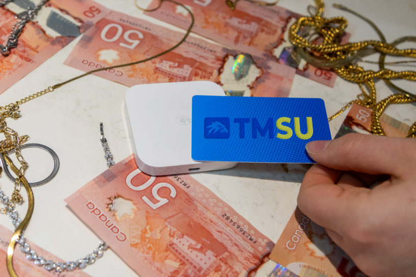 Image of someone tapping their card with a TMSU logo on it onto a card reader with $50 bills and gold chains around it.